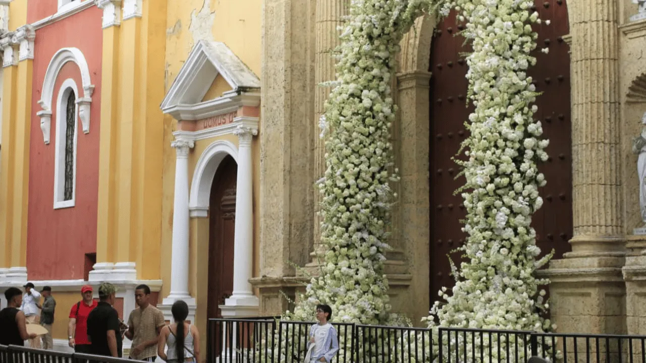 Arco de flores blancas sobre la puerta de madera de la catedral, con vallas y transeúntes.